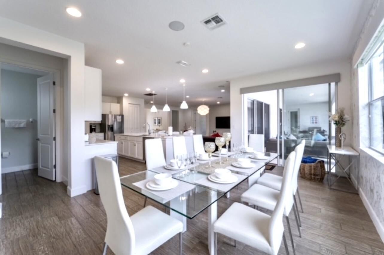 A modern dining area with a glass table, white chairs, and a kitchen in the background.