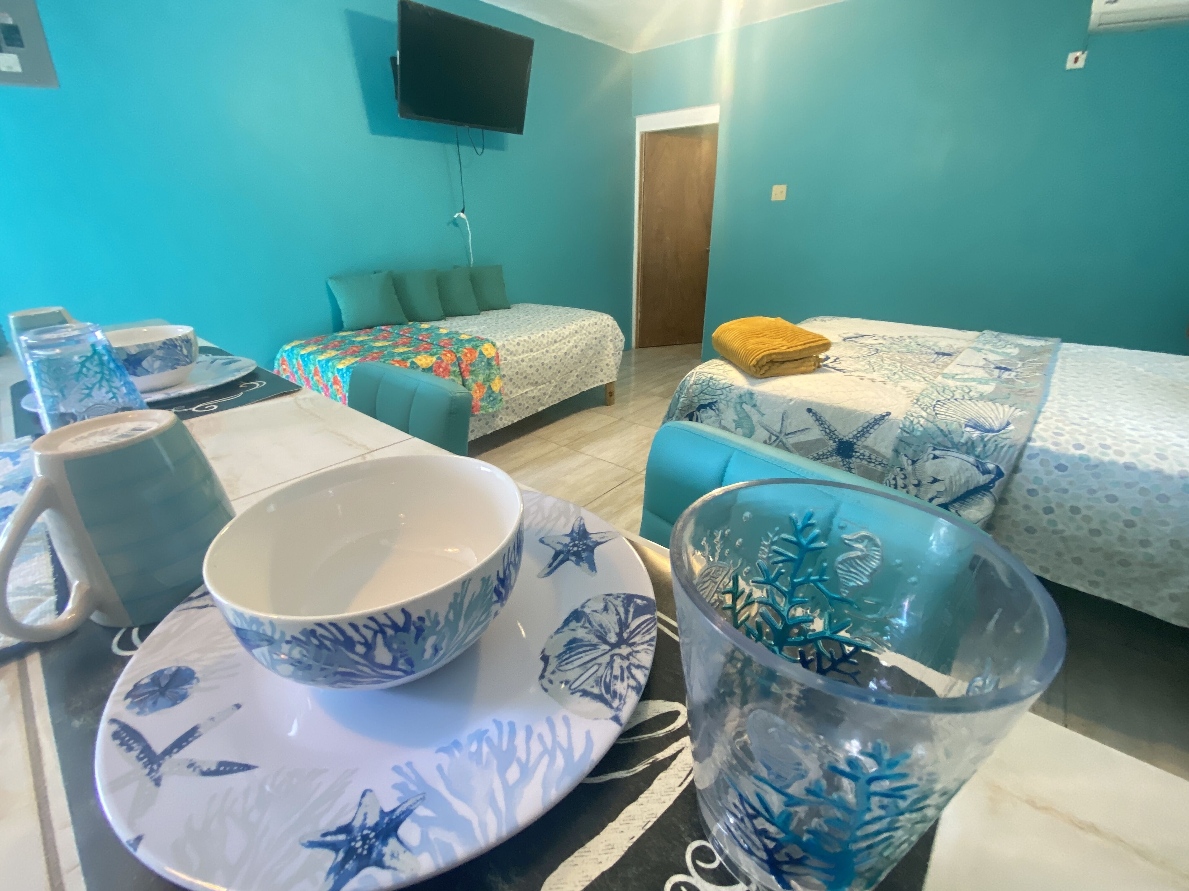 A room with two beds, a TV mounted on the wall, and a table with a bowl and a cup.
