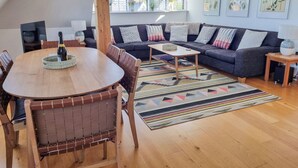 A modern living room with a dark-colored sofa, a wooden coffee table, and a dining table with chairs. There are framed botanical prints on the wall and a window with blinds. - Number 10 The Penthouse (Swanage)