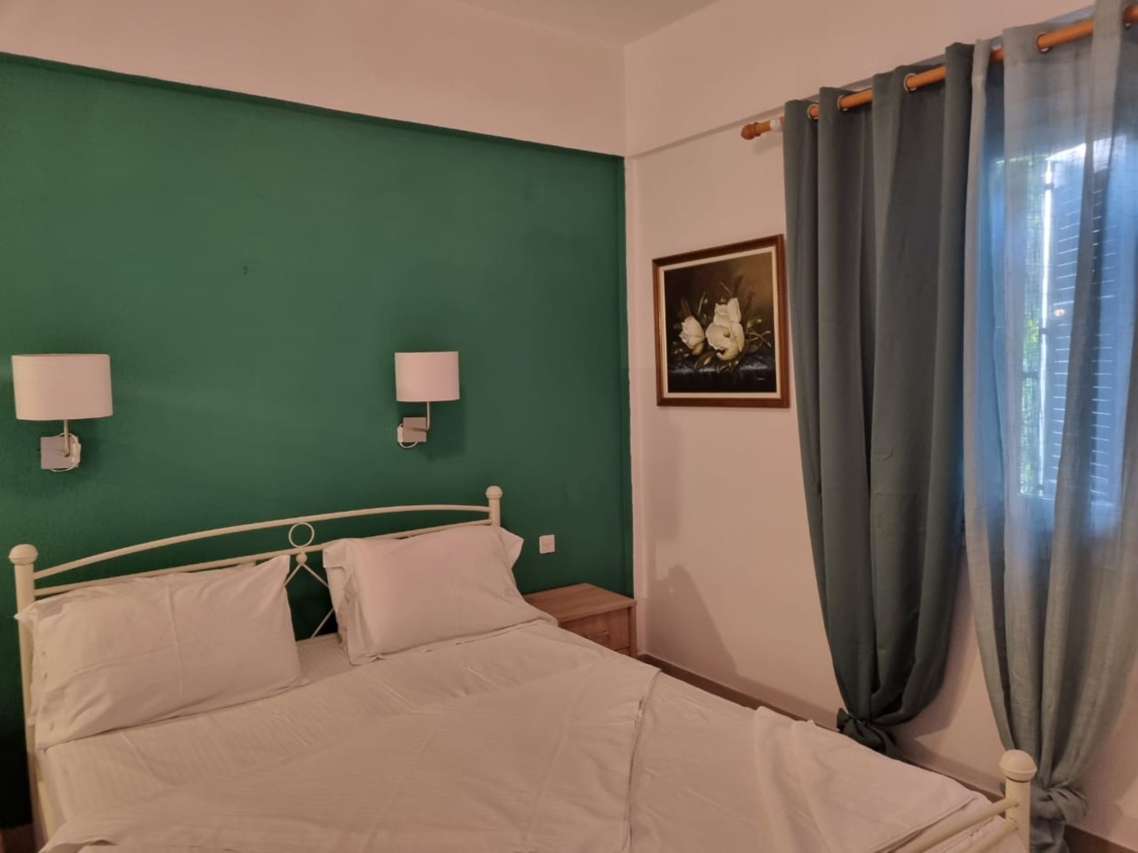 A bedroom with a green wall, a white bed, two bedside lamps, a framed picture, and a window with grey curtains.
