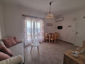 Un salon comprenant un canapé, une petite table, une table à manger avec des chaises, un ventilateur de plafond et une vue sur l’extérieur à travers une fenêtre ornée de rideaux à motifs floraux.