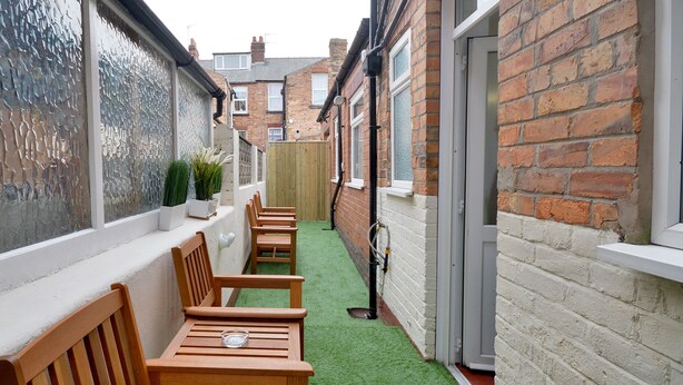A narrow outdoor patio with wooden benches and a green artificial turf floor, bordered by brick walls and a wooden fence. - Brightwater Ground Floor Apartment up to 6 Guests (Scarborough)