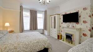 A bedroom with two beds, a television mounted above a fireplace, and a window with curtains. - Brightwater Ground Floor Apartment up to 6 Guests (Scarborough)