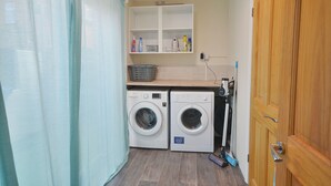A laundry room with two washing machines, a shelf with cleaning supplies, and a wooden door. - Brightwater Ground Floor Apartment up to 6 Guests (Scarborough)