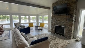 A spacious living room with a stone fireplace, a large TV mounted on the wall, and a view of a lake through large windows. - Beautiful lakefront property with hot tub  (Leesburg)