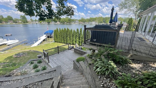 A wooden deck with a stone staircase leading to a covered area overlooking a lake, surrounded by greenery and a view of a dock with boats. - Beautiful lakefront property with hot tub  (Leesburg)