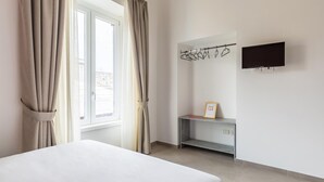 A hotel room with a bed, a TV mounted on the wall, a desk with a chair, and a window with curtains. - Maria s Bright Flat by Wonderful Italy - Duomo 2 (Naples)
