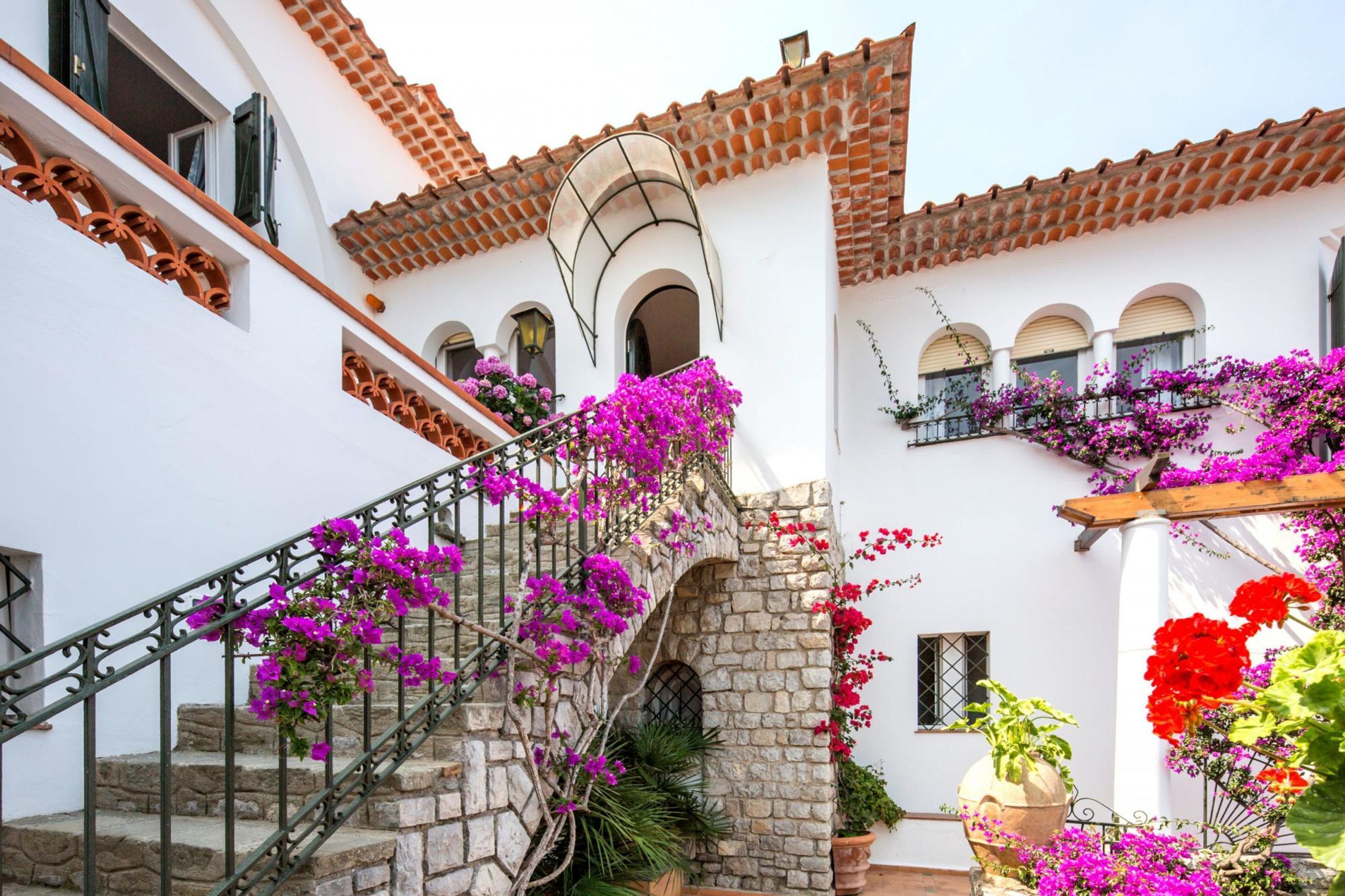 A white building with a stone staircase, a tiled roof, and balconies with flowers.