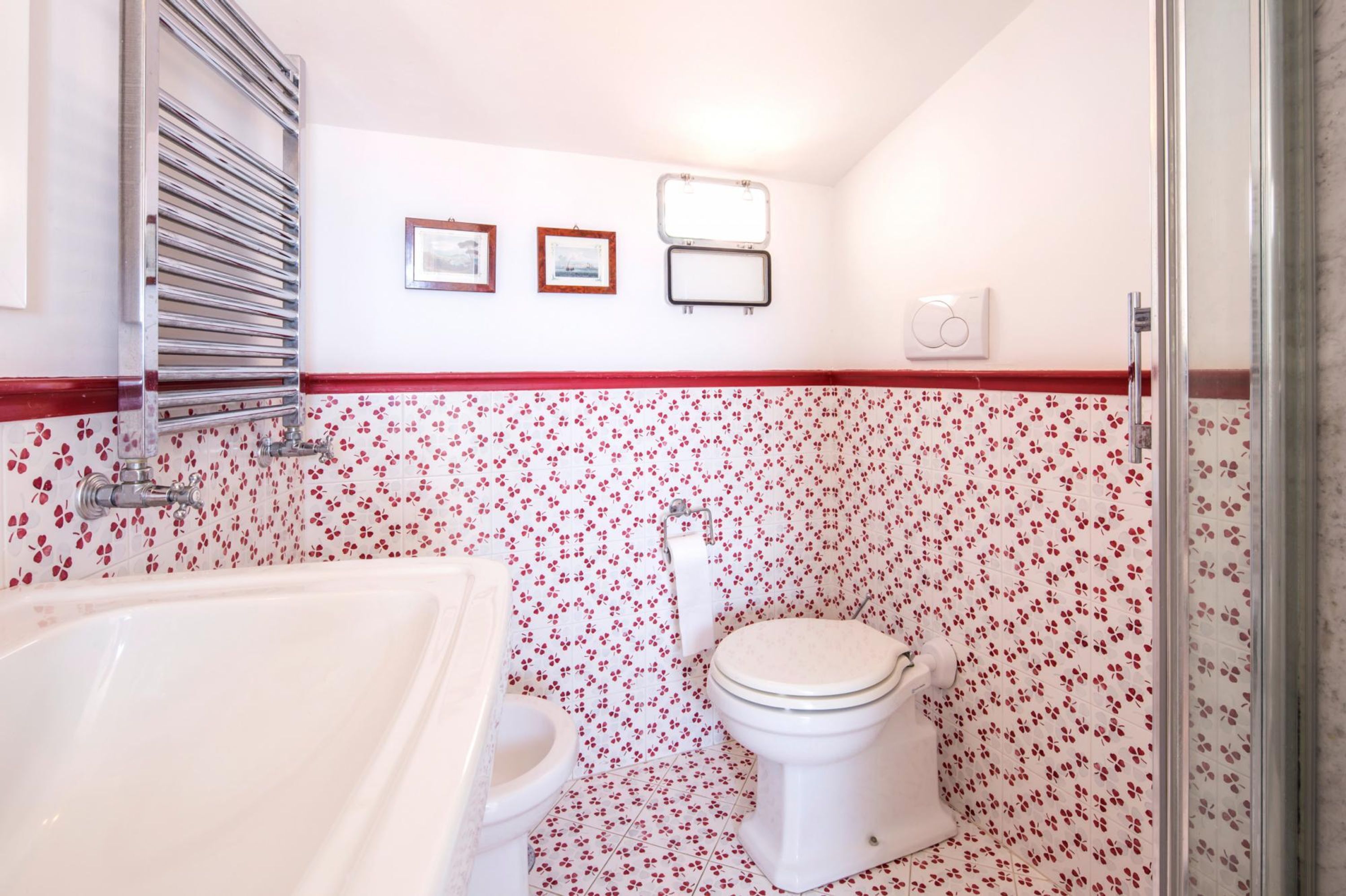 Um banheiro com banheira branca, vaso sanitário e chuveiro. As paredes têm um padrão floral em vermelho e branco. Há três quadros na parede.