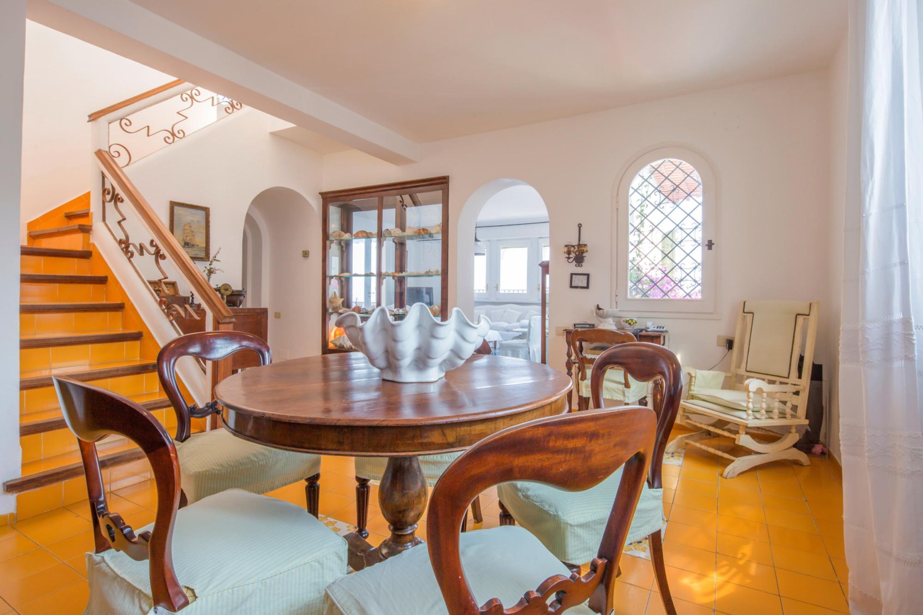 A dining area with a round wooden table, six chairs, and a decorative white pitcher. A staircase with wooden railings and a chandelier is visible in the background.