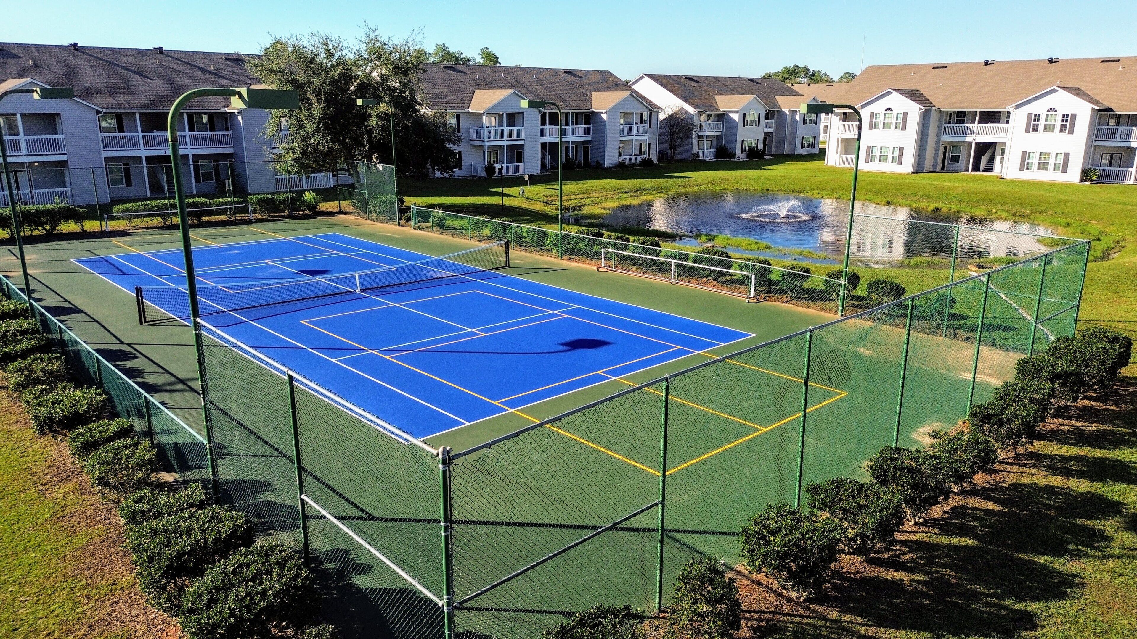 Een tennisbaan omringd door een groene hekken, met een klein vijvertje en woongebouwen op de achtergrond.