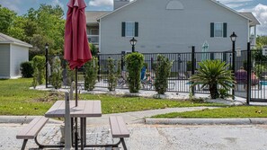 A picnic table with a red umbrella, a fenced pool area, and a white building in the background. - Updated, Ground Floor Unit w/ Pool & Gym, Sleeps 6 (Gulf Shores)