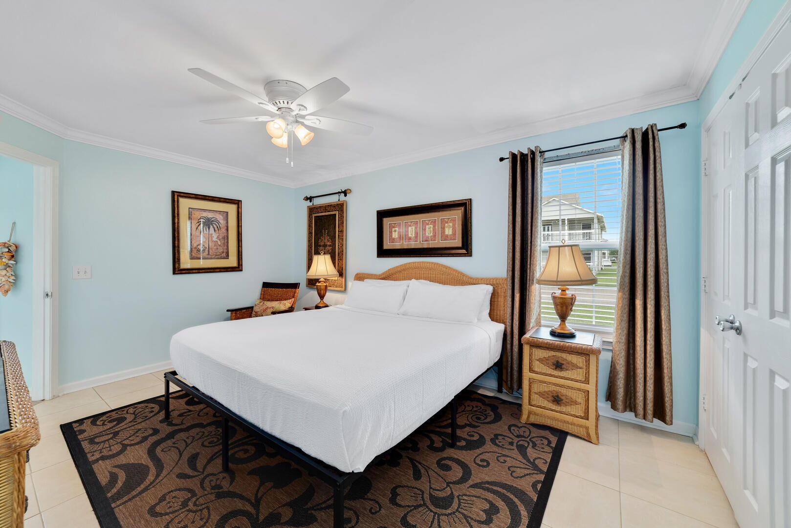 Habitación de hotel con cama, ventilador de techo, mesita de noche con lámpara, silla de mimbre y cuadros en las paredes.