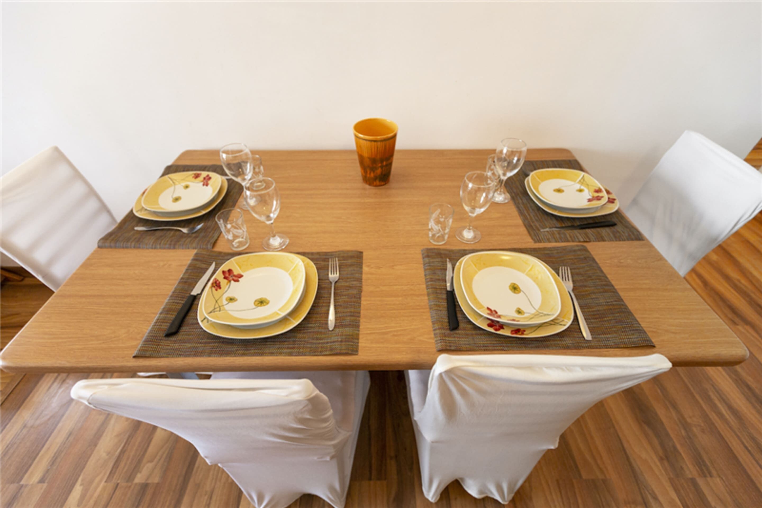 Mesa de comedor preparada para cuatro, con sillas blancas, una taza anaranjada en el centro y vajilla con motivos florales.