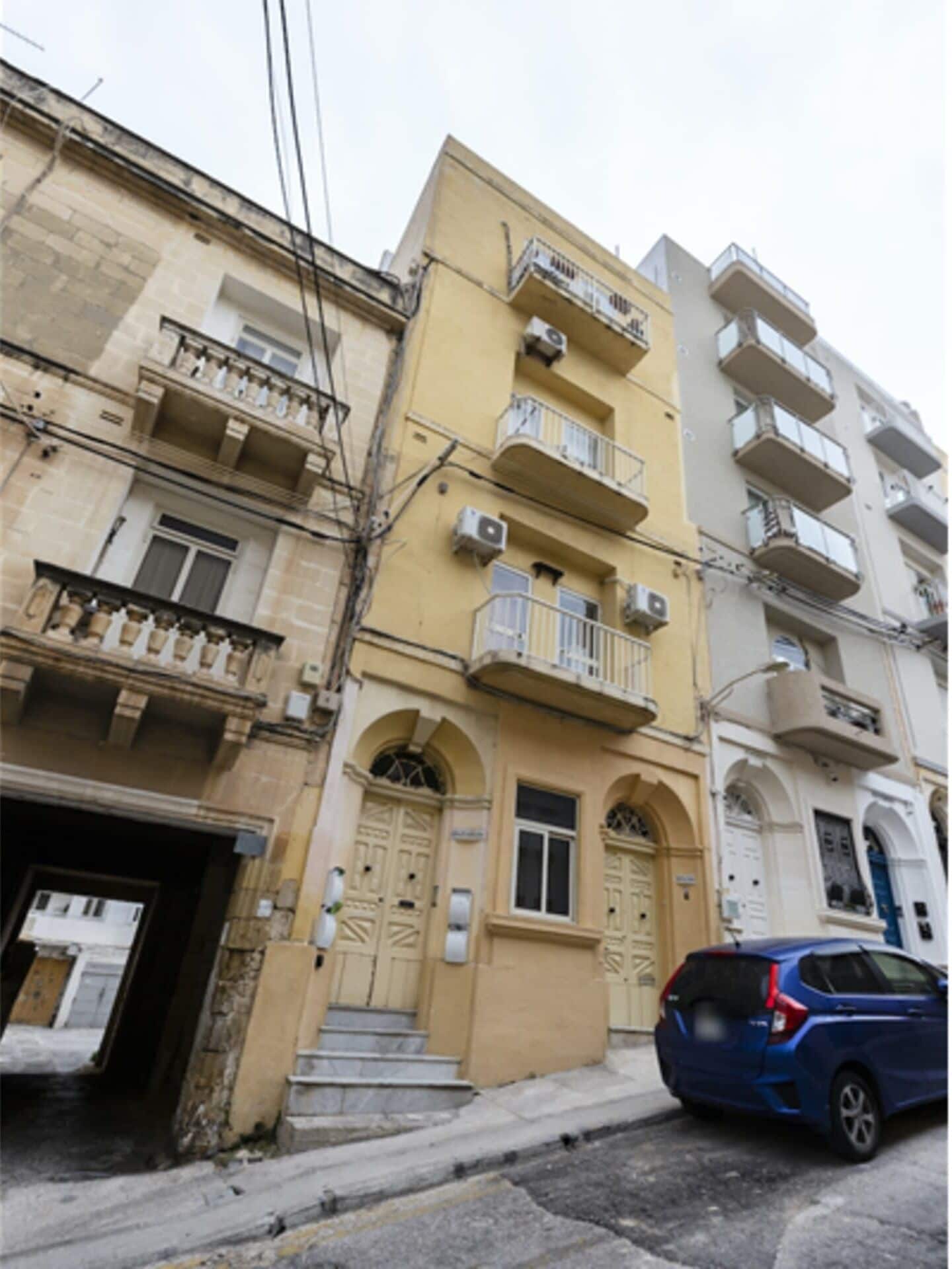 Un edificio de varios pisos con balcones, un coche estacionado al frente y una señal de calle a la vista.