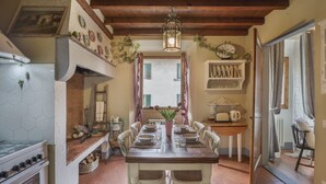 Un coin repas avec une table en bois dressée pour quatre personnes, entourée de chaises blanches. Un pot de fleurs est posé sur la table, et une fenêtre avec des rideaux se trouve derrière.