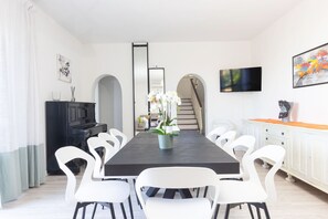 Un comedor moderno con una larga mesa, sillas blancas, una orquídea en maceta, un aparador negro y un televisor en la pared.