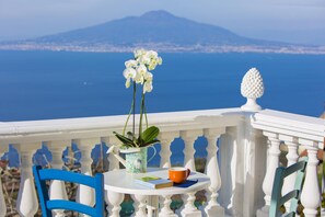 A white table with a blue chair, a potted plant, a book, and a mug on a balcony overlooking a body of water and a distant cityscape.