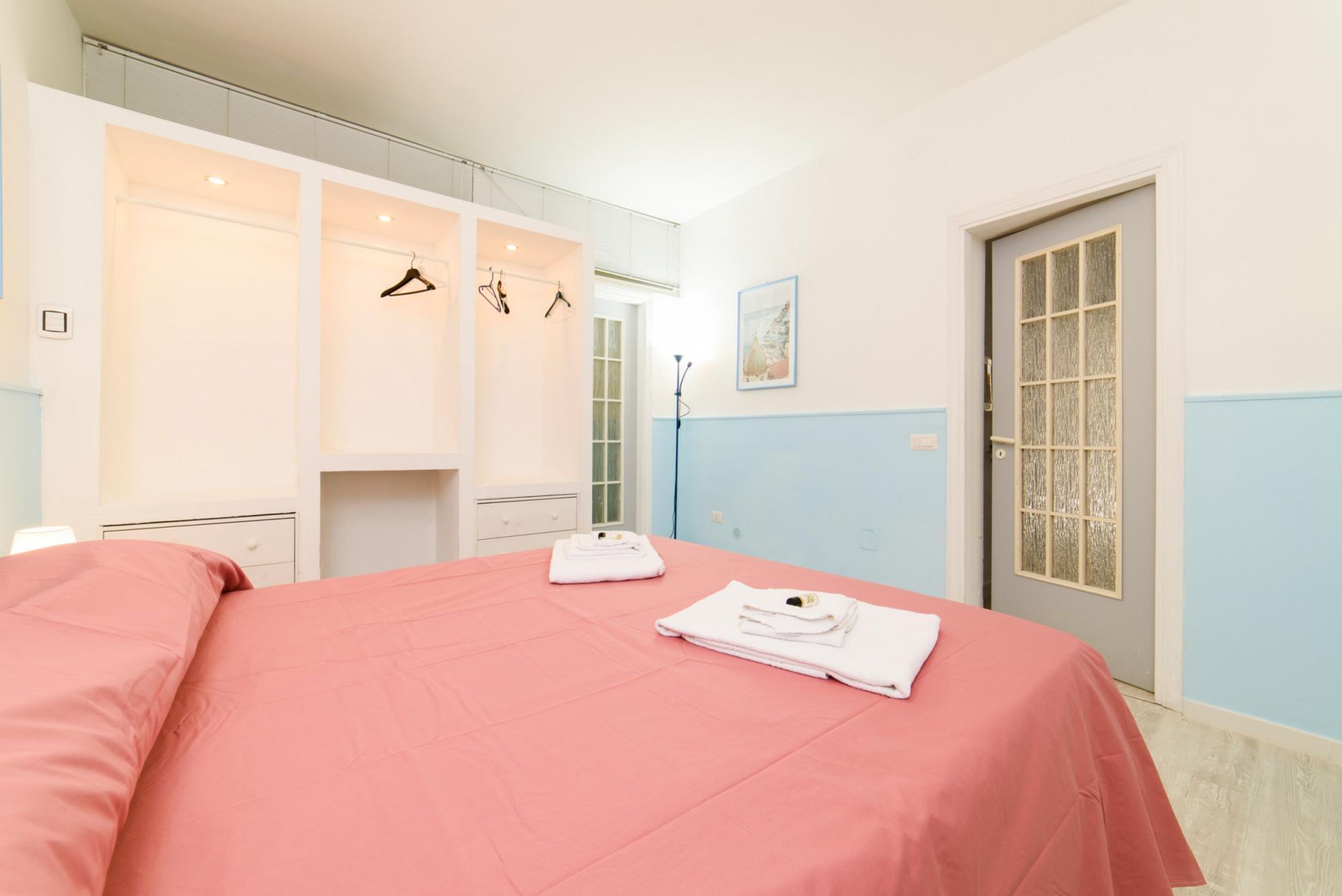 Quarto com colcha rosa, toalhas brancas e guarda-roupa com roupas penduradas.