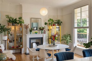 A well-lit room with a round dining table, blue chairs, a white fireplace, and a yellow cabinet with plants. - Archery Retreat (St Leonards-On-Sea)
