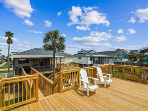 A wooden deck with white Adirondack chairs, a palm tree, and houses in the background under a clear sky.