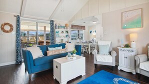 A spacious living room with a blue sofa, white armchairs, and a coffee table. There is a dining area with a table and chairs visible through a glass door. - Casa Hermosa by Ryson Vacation Rentals (Galveston)