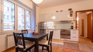 Una cocina compacta con armarios blancos, una mesa de comedor con sillas y vistas a la calle a través de amplias ventanas.