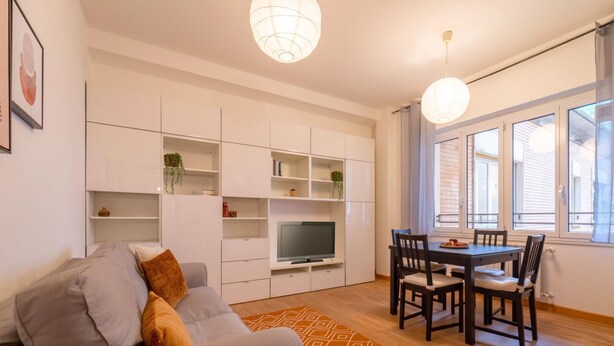 Una sala de estar moderna con sofá, televisor, mesa de comedor y sillas. Hay estanterías empotradas y una ventana con cortinas.