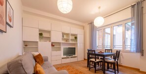 Una sala de estar moderna con sofá, televisor, mesa de comedor y sillas. Hay estanterías empotradas y una ventana con cortinas.