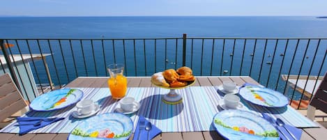 A table set for breakfast with plates, cutlery, and a fruit bowl, overlooking a body of water.