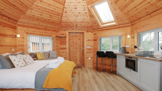L’intérieur chaleureux d’un chalet avec un plafond en bois, un lit recouvert d’une couverture jaune, un coin cuisine et une fenêtre donnant sur les arbres.