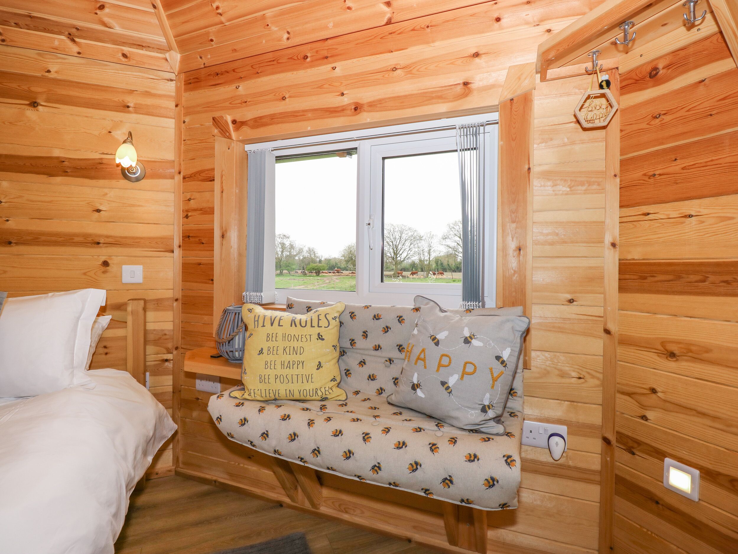 Acogedor cuarto de cabaña de madera con una cama, un banco con cojines con motivos de abejas y una ventana con vistas al exterior.