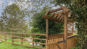 Un pont en bois, surmonté d’une grille métallique, entouré de verdure et bordé par une étendue d’eau.