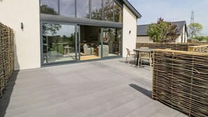 A modern house with large glass windows, a patio area with outdoor furniture, and a clear view of the surrounding landscape. - The Barn (Amlwch)