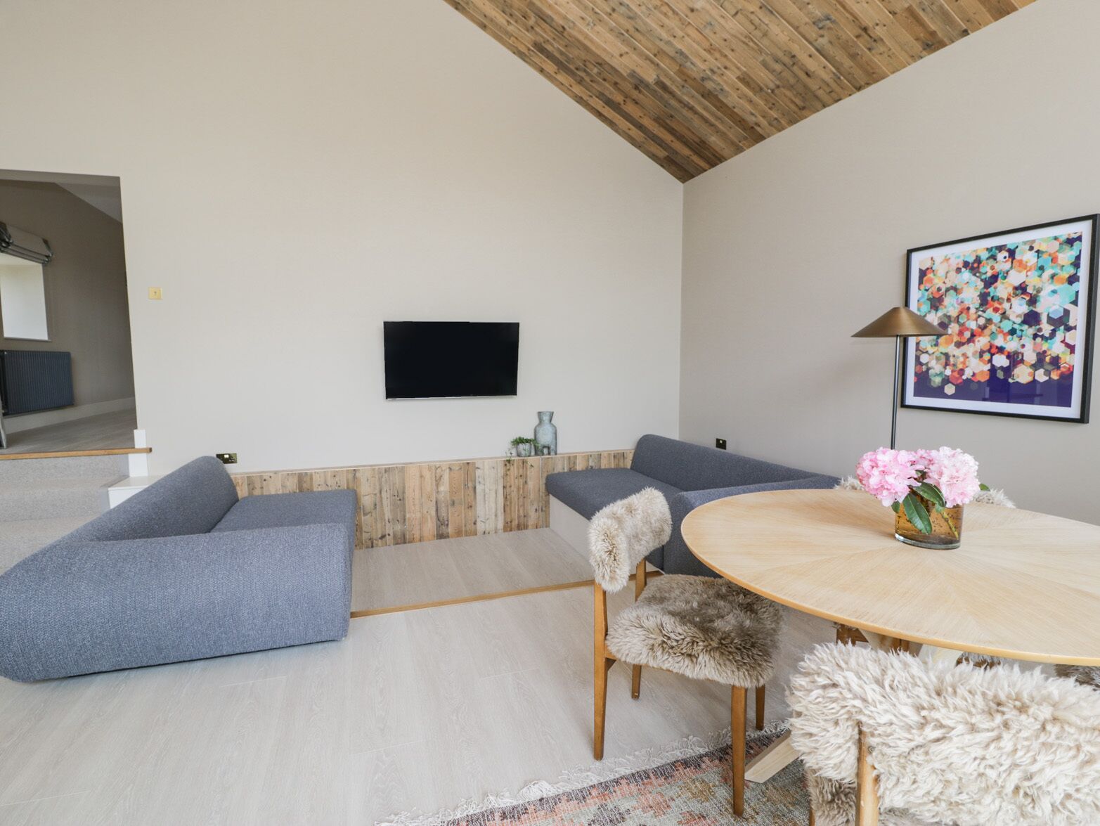 En moderne stue med træloft, et fladskærms-tv på væggen, et rundt træbord med en vase med blomster og en sofa med bløde puder.