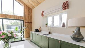A kitchen with green cabinets, a white countertop, a window with blinds, and a vase with flowers on the counter. - The Barn (Amlwch)
