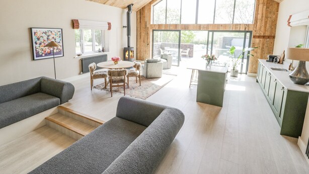A spacious living area with a high ceiling, a fireplace, a dining table with chairs, and a kitchen area. - The Barn (Amlwch)