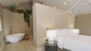 A modern bedroom with a large bed, a freestanding bathtub, and a bedside table with a lamp. - The Barn (Amlwch)