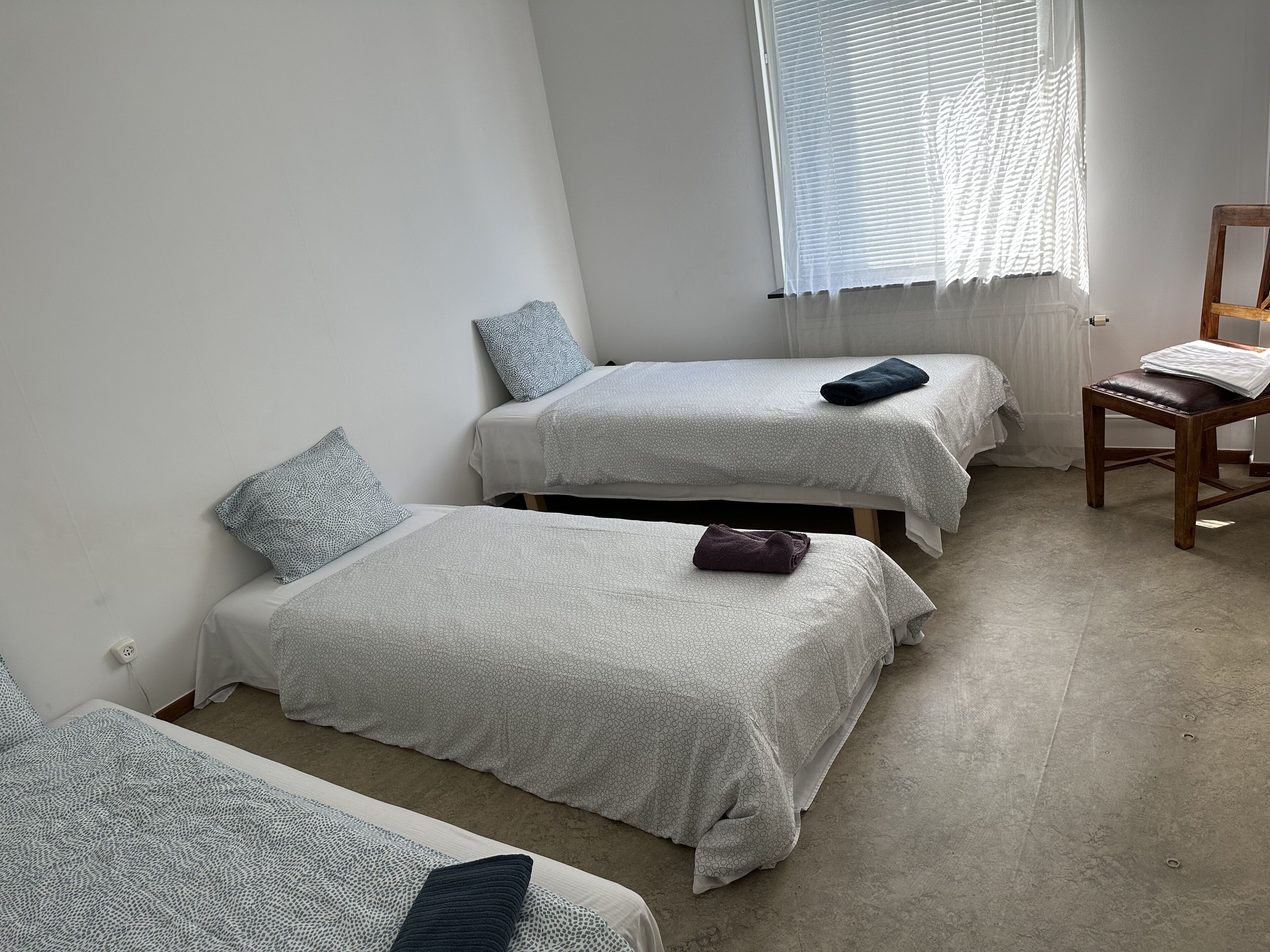 A room with three beds, each with a patterned bedspread and pillows, a wooden chair, and a window with blinds.