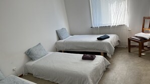 Habitación con tres camas, cada una con edredón y almohadas estampadas, una silla de madera y una ventana con persianas.