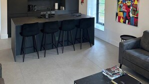 Een moderne keuken met een centraal eiland en barstoelen, een zithoek met een bank en salontafel, en een kleurrijk abstract schilderij aan de muur.