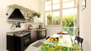 Uma cozinha compacta com fogão a gás, forno e exaustor preto. Uma mesa de jantar com toalha de mesa florida e cadeiras está montada perto de uma janela grande com vista para vegetação.