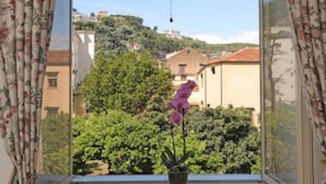 Uma janela com cortinas florais, um brinquedo pendurado e um vaso com flores cor-de-rosa.