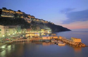 A coastal town at dusk with buildings on a hillside and boats docked at the waterfront. - Casa Chiarina in Sorrento (Sorrento)