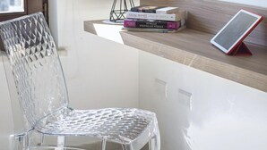 Una silla de vidrio transparente, un escritorio con una lámpara y libros en una estantería, todo en una habitación con ventana.