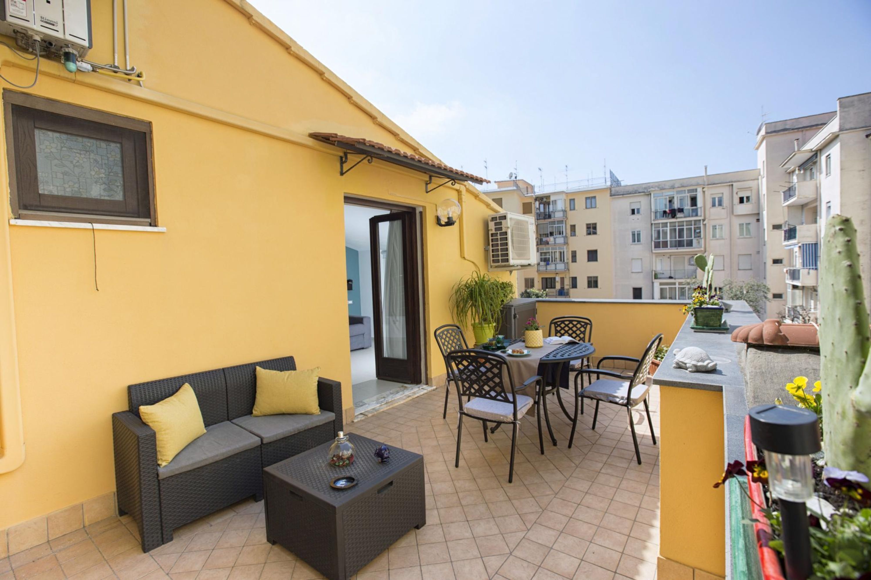 A yellow building with a balcony featuring a sofa, chairs, a table, and potted plants.