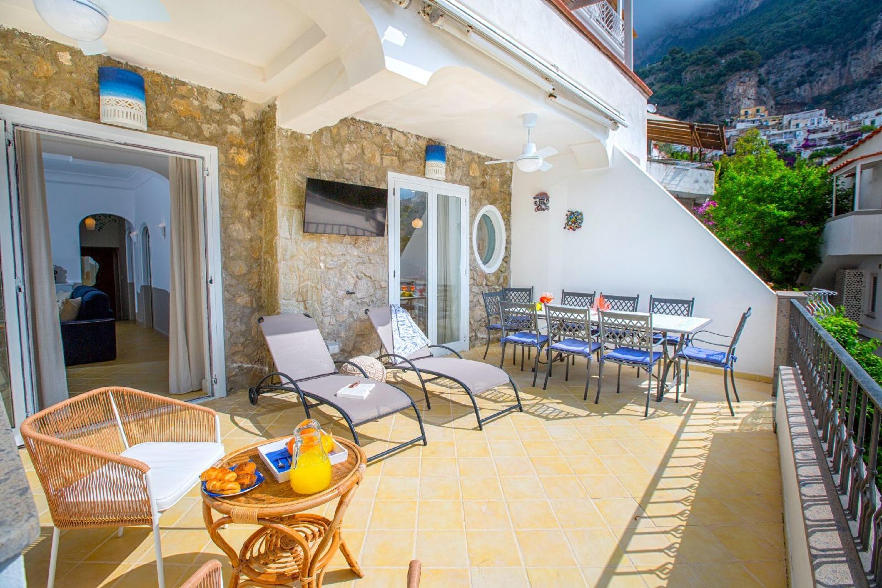 Un patio al aire libre bañado por el sol, con mobiliario de mimbre, una mesa con cesta de fruta y vistas a un pueblo montañoso.