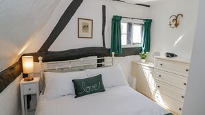 A bedroom with a white bed, a nightstand with a lamp, a dresser, a window with green curtains, and a picture on the wall. - Bluebell Cottage (Stratford-upon-Avon)