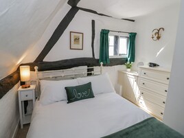 A bedroom with a white bed, a nightstand with a lamp, a dresser, a window with green curtains, and a picture on the wall.
