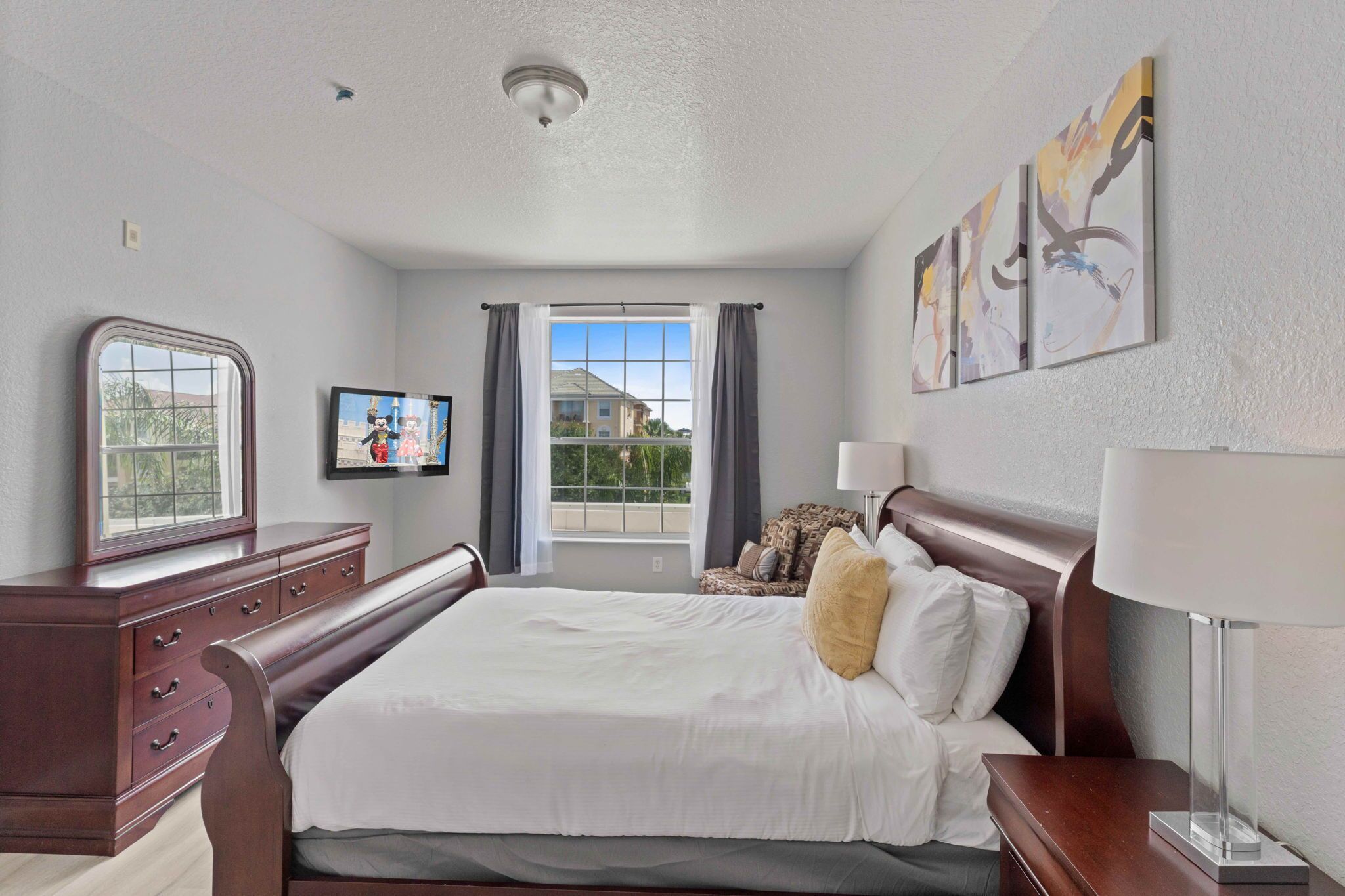 A hotel room with a large bed, a dresser, a window with curtains, a television, and artwork on the wall.